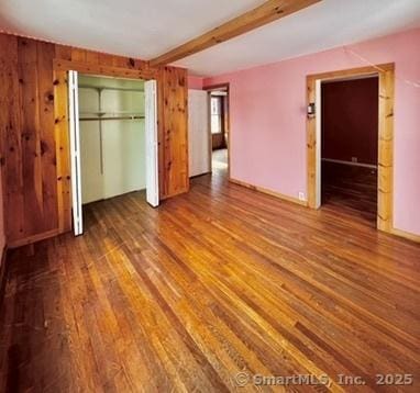 unfurnished bedroom with beamed ceiling, wooden walls, wood finished floors, and a closet