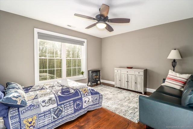 bedroom featuring a wood stove, a ceiling fan, visible vents, baseboards, and dark wood-style flooring
