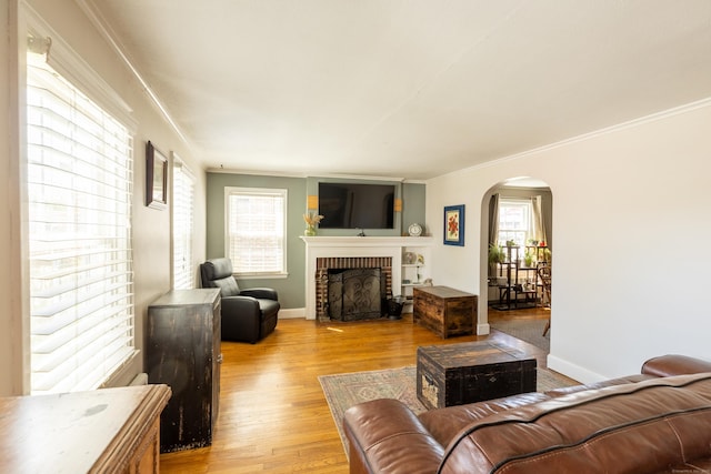 living room with crown molding, a brick fireplace, arched walkways, light wood-style flooring, and baseboards