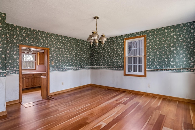 empty room with wallpapered walls, a wainscoted wall, an inviting chandelier, light wood-type flooring, and a textured ceiling