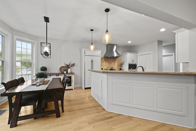 kitchen with built in appliances, light wood-style flooring, decorative light fixtures, wall chimney range hood, and white cabinets