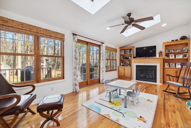 living area featuring wood finished floors, vaulted ceiling with skylight, ceiling fan, a glass covered fireplace, and baseboards