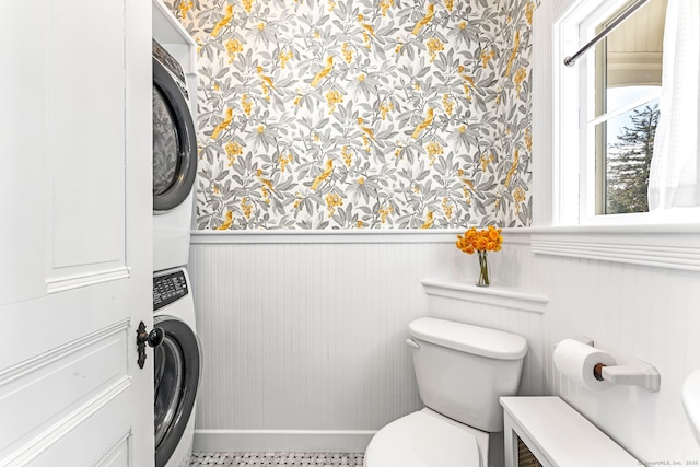 bathroom with wainscoting, toilet, baseboards, stacked washing maching and dryer, and wallpapered walls