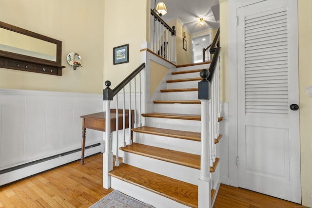 staircase featuring a baseboard heating unit, a towering ceiling, a wainscoted wall, and wood finished floors