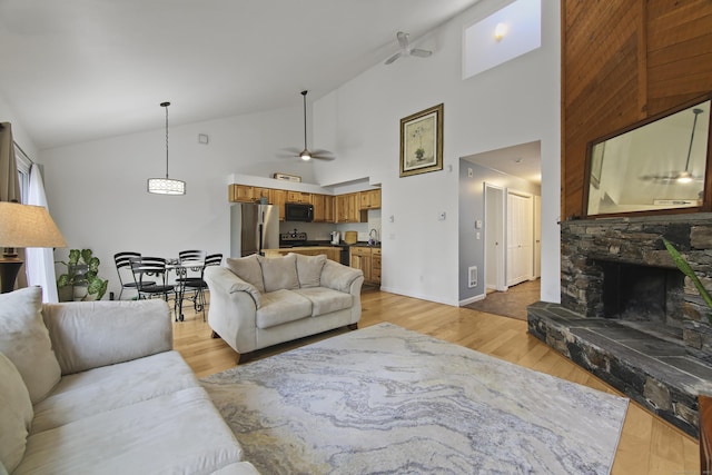 living area featuring ceiling fan, baseboards, light wood-type flooring, high vaulted ceiling, and a stone fireplace