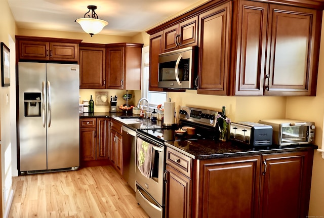 kitchen with dark stone counters, light wood-style flooring, a sink, appliances with stainless steel finishes, and hanging light fixtures