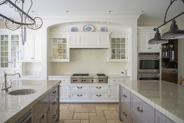 kitchen with appliances with stainless steel finishes, a sink, stone tile flooring, hanging light fixtures, and glass insert cabinets