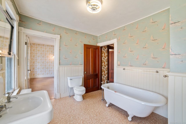 bathroom featuring a sink, wallpapered walls, toilet, a freestanding tub, and wainscoting