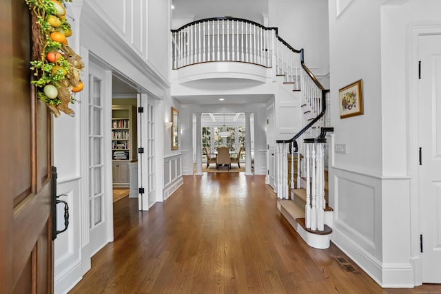 foyer entrance featuring stairs, a decorative wall, a wainscoted wall, dark wood-style flooring, and a towering ceiling