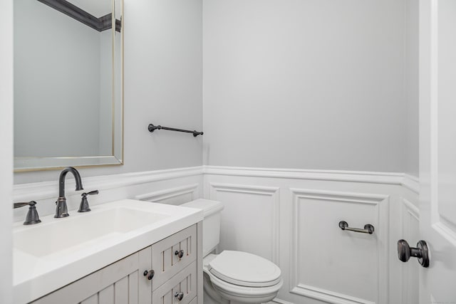 half bath with vanity, toilet, wainscoting, and a decorative wall