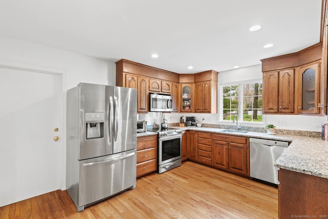 kitchen featuring glass insert cabinets, brown cabinets, light stone counters, a sink, and appliances with stainless steel finishes