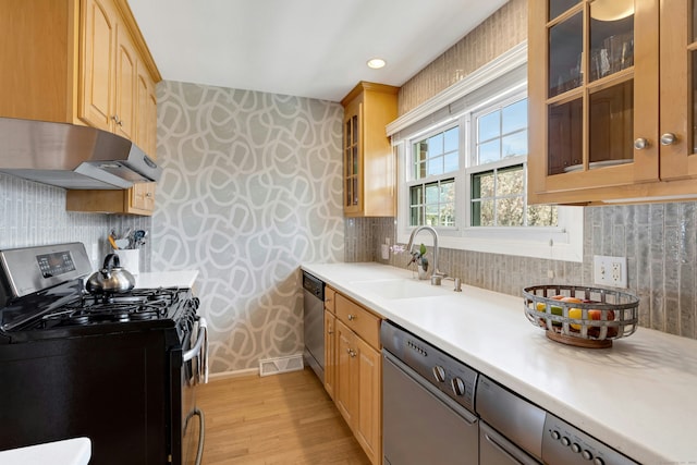 kitchen featuring appliances with stainless steel finishes, light wood-type flooring, a sink, under cabinet range hood, and wallpapered walls