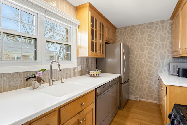 kitchen featuring appliances with stainless steel finishes, a sink, glass insert cabinets, wallpapered walls, and light wood-style flooring