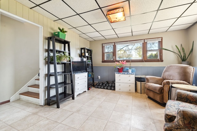 living area with light tile patterned flooring, wainscoting, wood walls, stairs, and a drop ceiling