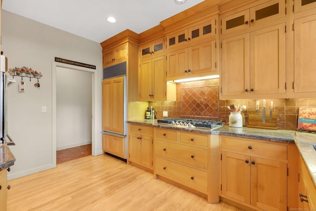 kitchen with dark stone countertops, glass insert cabinets, decorative backsplash, paneled built in fridge, and light wood-type flooring