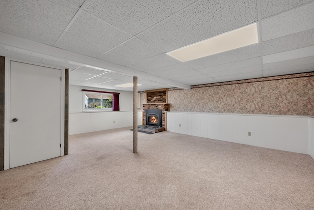 basement featuring a wainscoted wall, a wood stove, a drop ceiling, and carpet flooring