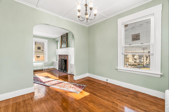 unfurnished living room with radiator heating unit, arched walkways, baseboards, a brick fireplace, and hardwood / wood-style flooring