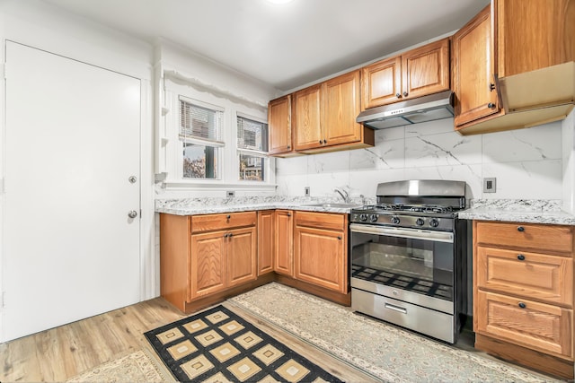 kitchen with a sink, stainless steel gas range, decorative backsplash, light stone countertops, and under cabinet range hood