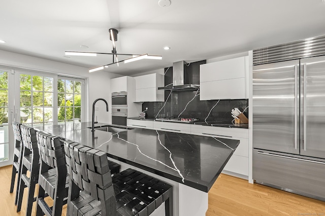 kitchen with modern cabinets, wall chimney range hood, a large island with sink, a sink, and appliances with stainless steel finishes