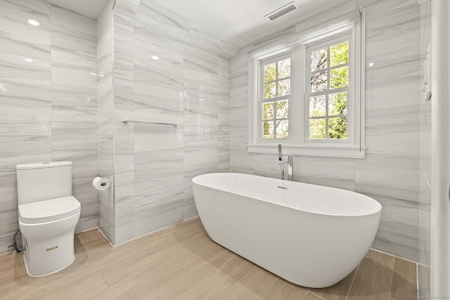bathroom featuring toilet, a freestanding tub, tile walls, and visible vents