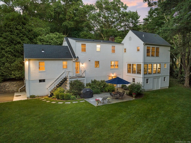 back of property at dusk with a patio, stairway, a shingled roof, a lawn, and a chimney