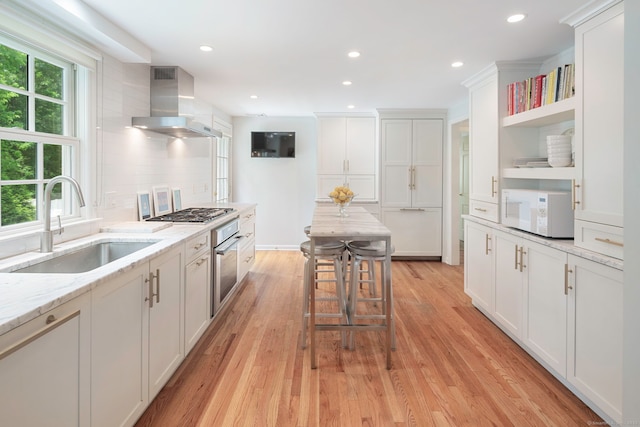 kitchen featuring wall chimney range hood, open shelves, white cabinets, a sink, and appliances with stainless steel finishes