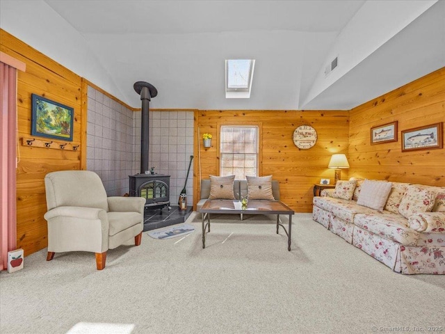 living room featuring a wood stove, vaulted ceiling with skylight, visible vents, carpet floors, and wooden walls