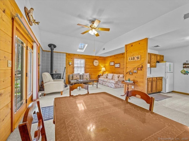 dining area featuring a wood stove, light colored carpet, wooden walls, a ceiling fan, and a skylight