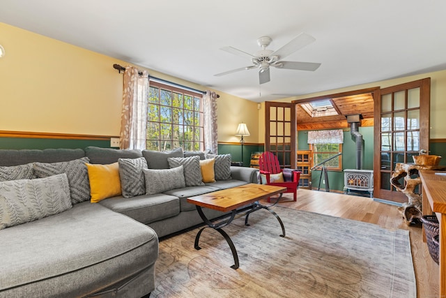 living room featuring a wood stove, wood finished floors, wainscoting, a ceiling fan, and a skylight