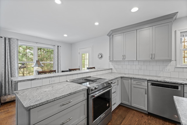 kitchen with stainless steel appliances, a peninsula, gray cabinets, dark wood-style flooring, and light stone countertops
