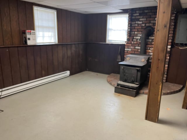 basement featuring a wood stove, a baseboard radiator, electric panel, wooden walls, and a paneled ceiling