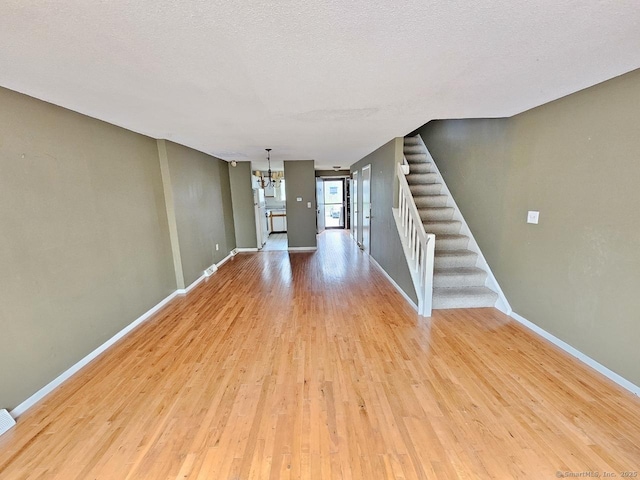 unfurnished living room with an inviting chandelier, light wood-style flooring, baseboards, stairway, and a textured ceiling