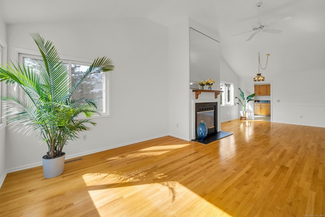 unfurnished living room with high vaulted ceiling, ceiling fan, a glass covered fireplace, light wood-style flooring, and baseboards
