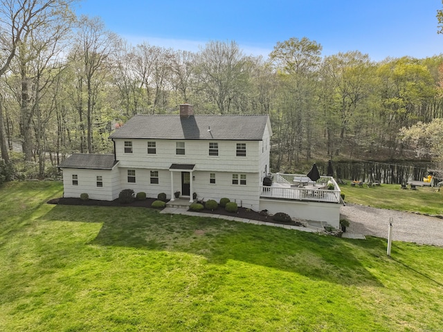 view of front of property with a front yard, a deck, a chimney, a shingled roof, and a view of trees