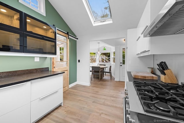 kitchen with stainless steel gas stove, light wood-style floors, lofted ceiling with skylight, wall chimney range hood, and white cabinetry
