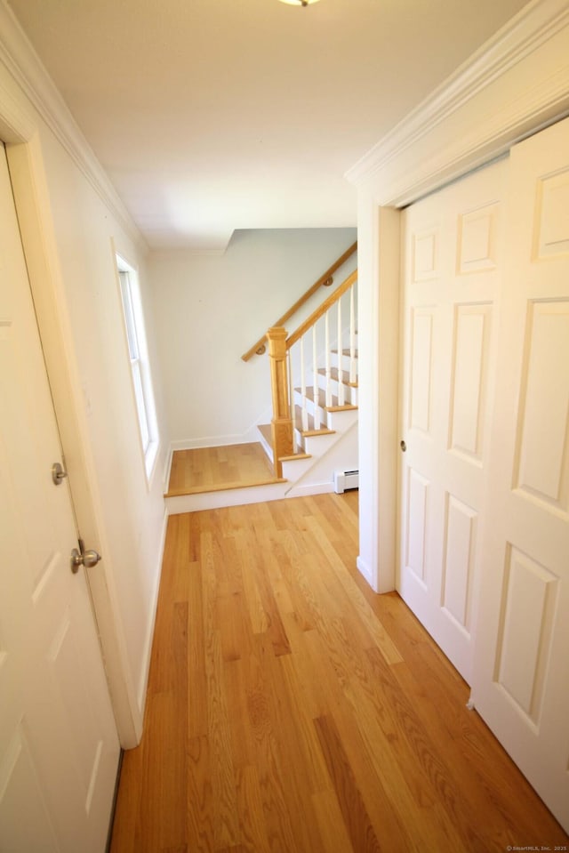 corridor featuring a baseboard radiator, baseboards, light wood-type flooring, ornamental molding, and stairway