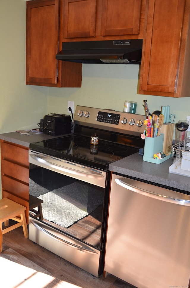 kitchen with stainless steel appliances, under cabinet range hood, brown cabinetry, dark countertops, and wood finished floors