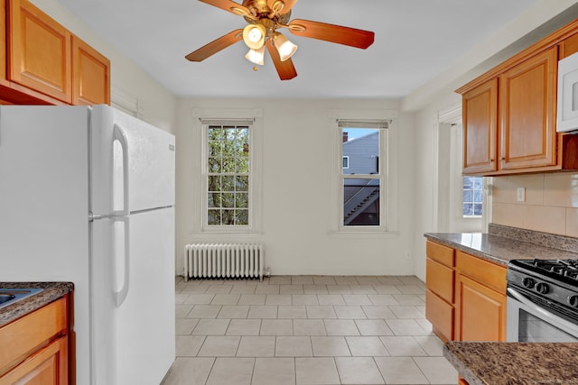 kitchen with dark stone countertops, decorative backsplash, ceiling fan, radiator heating unit, and white appliances