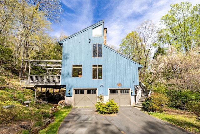 view of side of property with aphalt driveway, a wooden deck, a chimney, stairs, and an attached garage