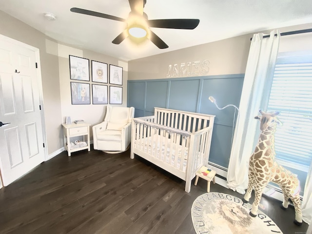 bedroom with wood finished floors, a decorative wall, ceiling fan, a nursery area, and a baseboard radiator