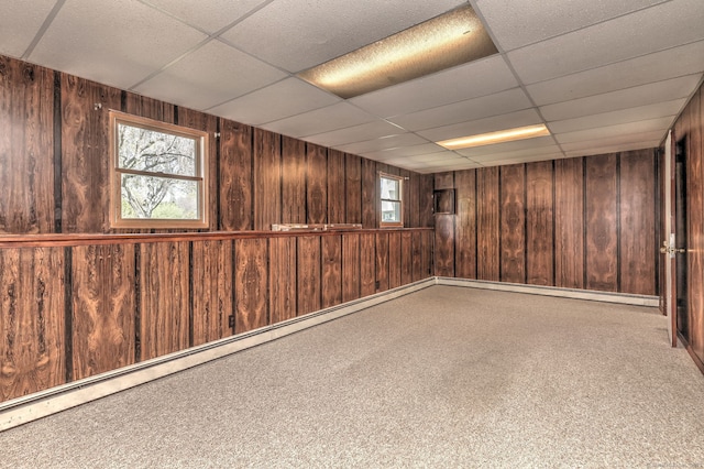 basement featuring a baseboard radiator, wooden walls, a healthy amount of sunlight, and a paneled ceiling