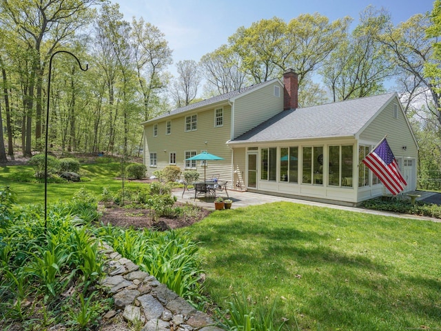 rear view of property featuring a chimney, a patio, a sunroom, a lawn, and a shingled roof