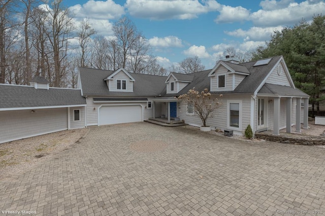cape cod home featuring an attached garage, a shingled roof, roof mounted solar panels, a chimney, and decorative driveway