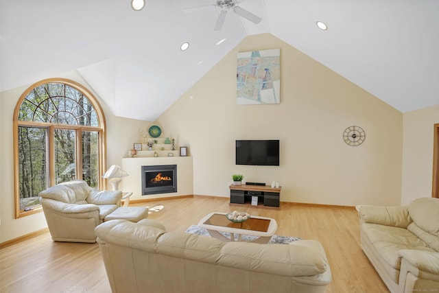 living room with a warm lit fireplace, ceiling fan, baseboards, light wood-type flooring, and vaulted ceiling