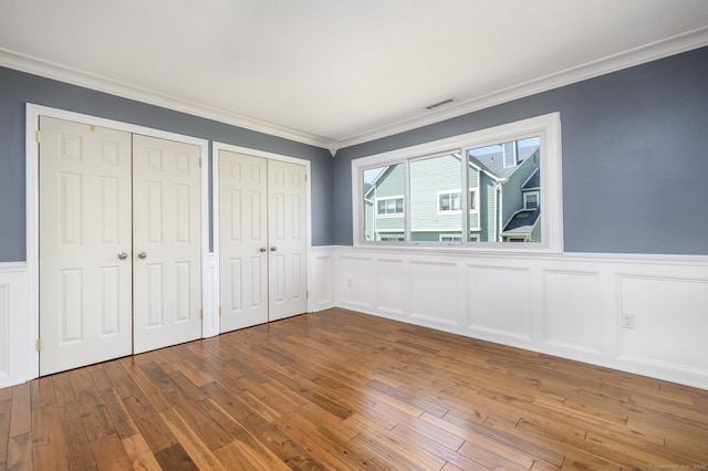 unfurnished bedroom featuring a wainscoted wall, multiple closets, crown molding, visible vents, and hardwood / wood-style flooring