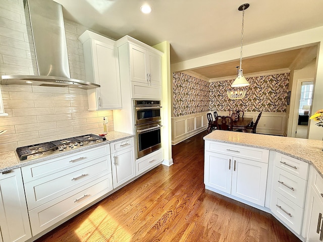 kitchen with light wood-style floors, wall chimney exhaust hood, white cabinetry, decorative light fixtures, and stainless steel appliances