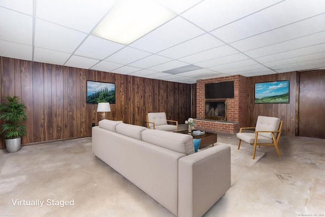 living room with a paneled ceiling, a brick fireplace, wood walls, and concrete flooring