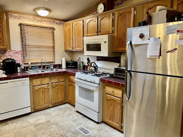 kitchen featuring a sink, brown cabinetry, white appliances, a textured ceiling, and tile countertops