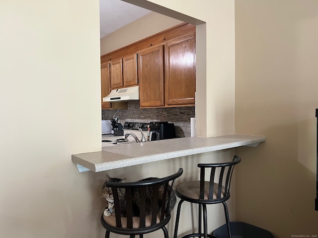 kitchen with brown cabinets, electric stove, decorative backsplash, a kitchen bar, and under cabinet range hood