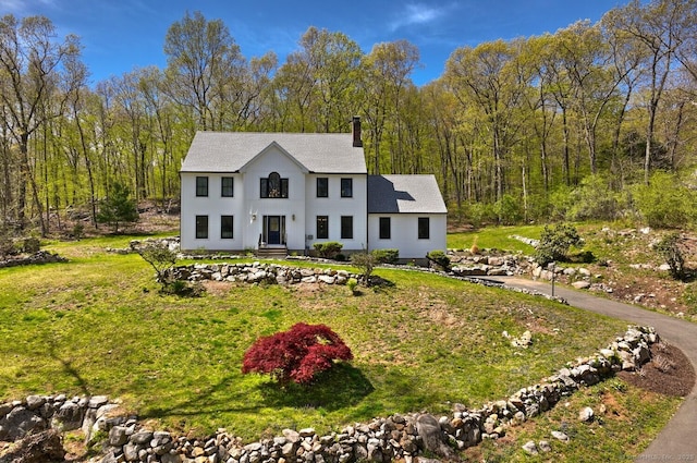 view of front of home with a chimney, a shingled roof, a wooded view, and a front yard